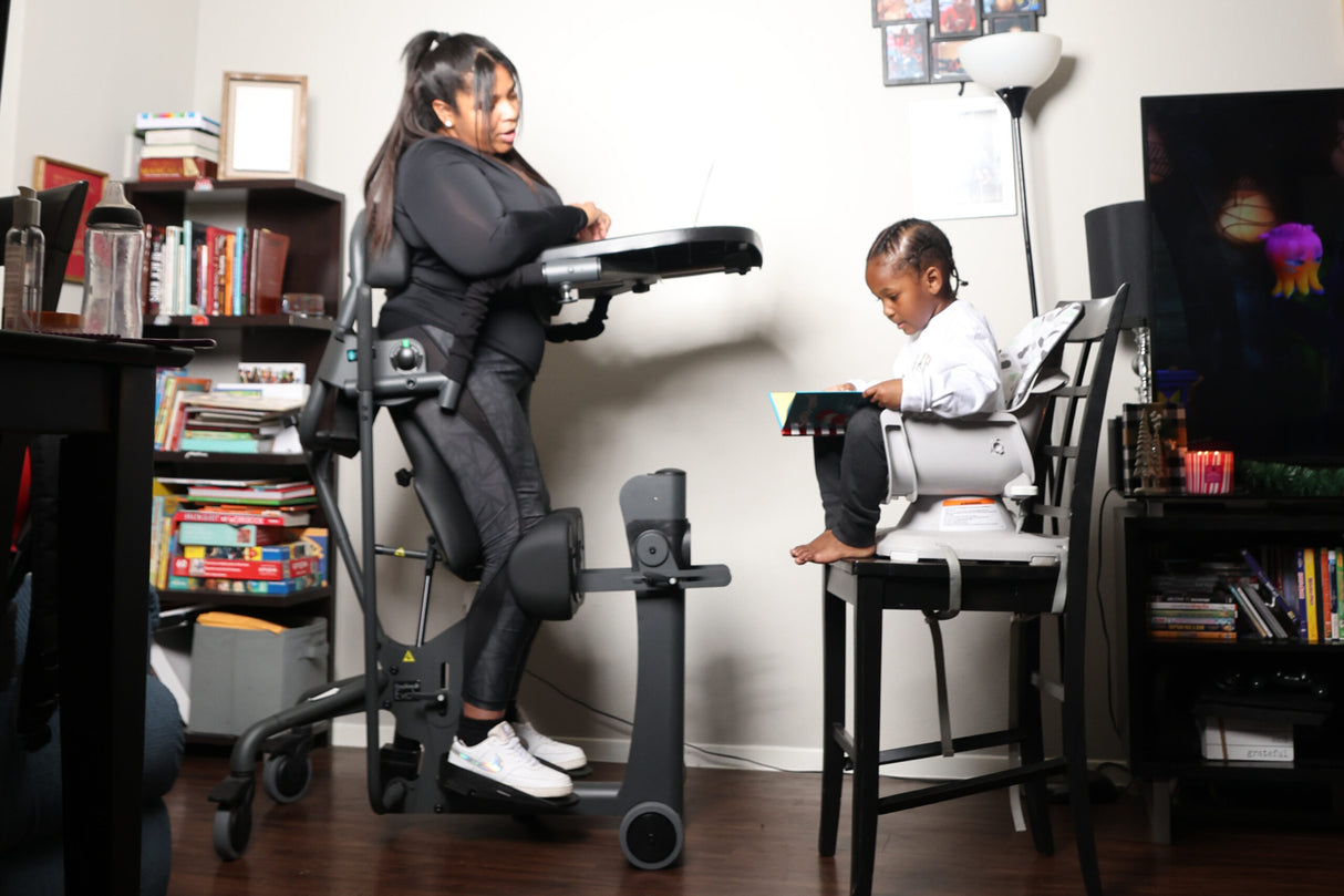 A woman uses an EasyStand Evolv office environment, with a young child seated in a white high chair facing her. The room contains bookshelves, a desk, and a television.