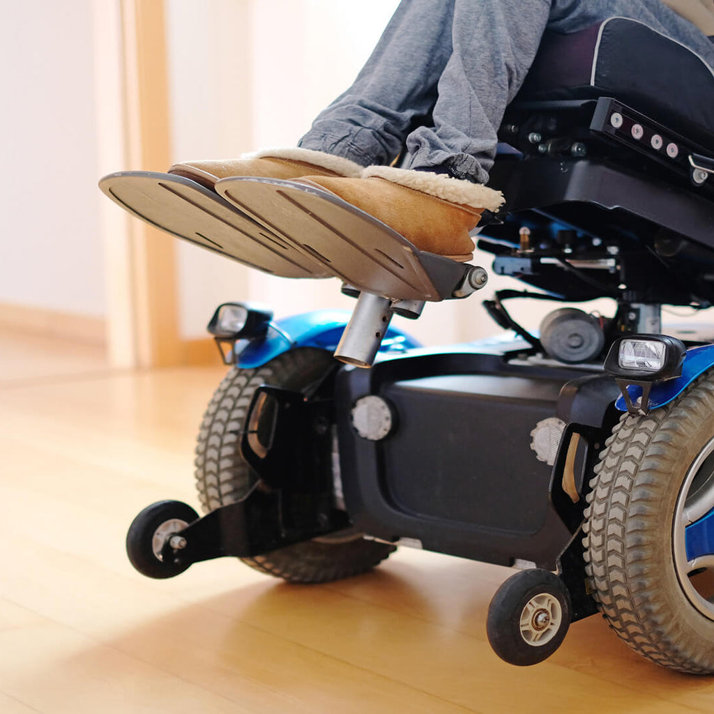 the base of a blue and black power wheelchair is viewed from close to the floor. The chair is slightly tilted and is sitting on a light hardwood floor in a well-lit room.