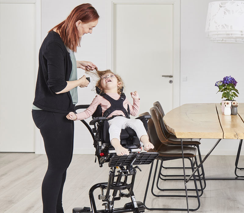 Woman assisting a child in an XO Frame High-Low Chair at a dining table in a home setting