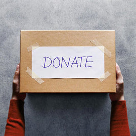 a pair of arms wearing a red sweater hold out a cardboard box over a concrete background. a white sheet of paper with the word 'donate' in purple block letters is taped to the top of the box