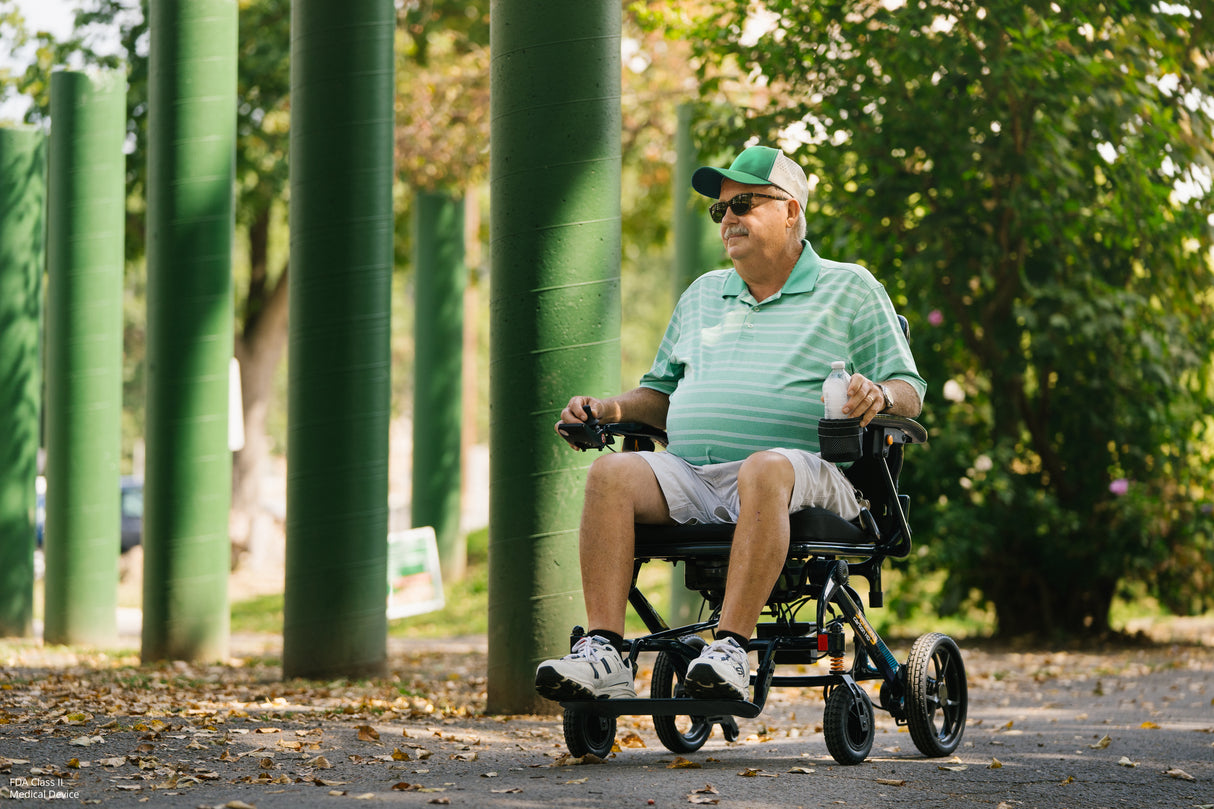 Man in a green shirt and cap using a Jazzy Carbon HD Power Chair in a park setting with trees and green pillars.