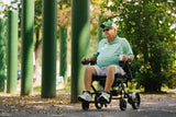 Man in a green shirt and cap using a Jazzy Carbon HD Power Chair in a park setting with trees and green pillars.