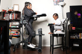 A woman uses an EasyStand Evolv office environment, with a young child seated in a white high chair facing her. The room contains bookshelves, a desk, and a television.