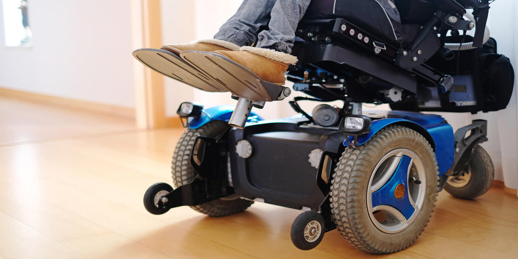 the base of a blue and black power wheelchair is viewed from close to the floor. The chair is slightly tilted and is sitting on a light hardwood floor in a well-lit room.