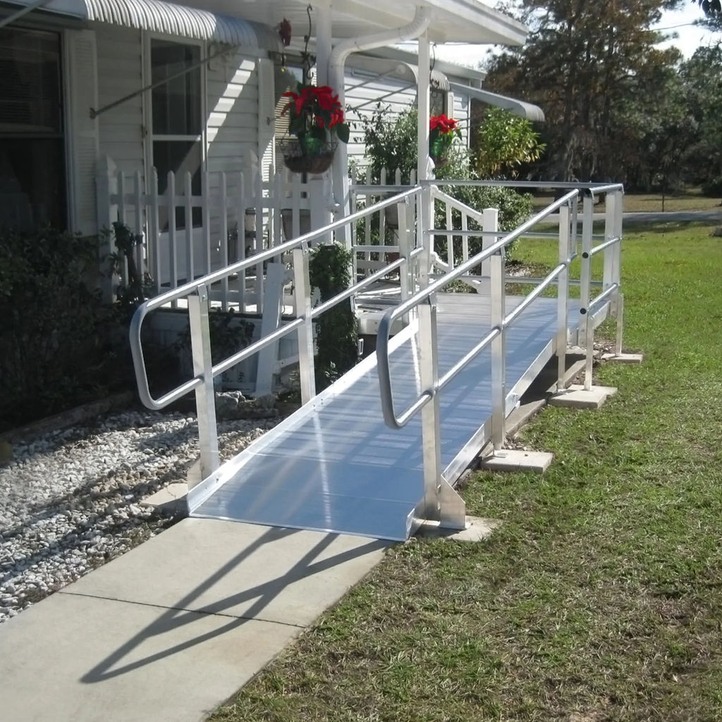 a silver aluminum wheelchair ramp is installed on the front of a white house with an awning. the ramp has a flat platform at the top and make a turn to lead to the concrete walkway below.