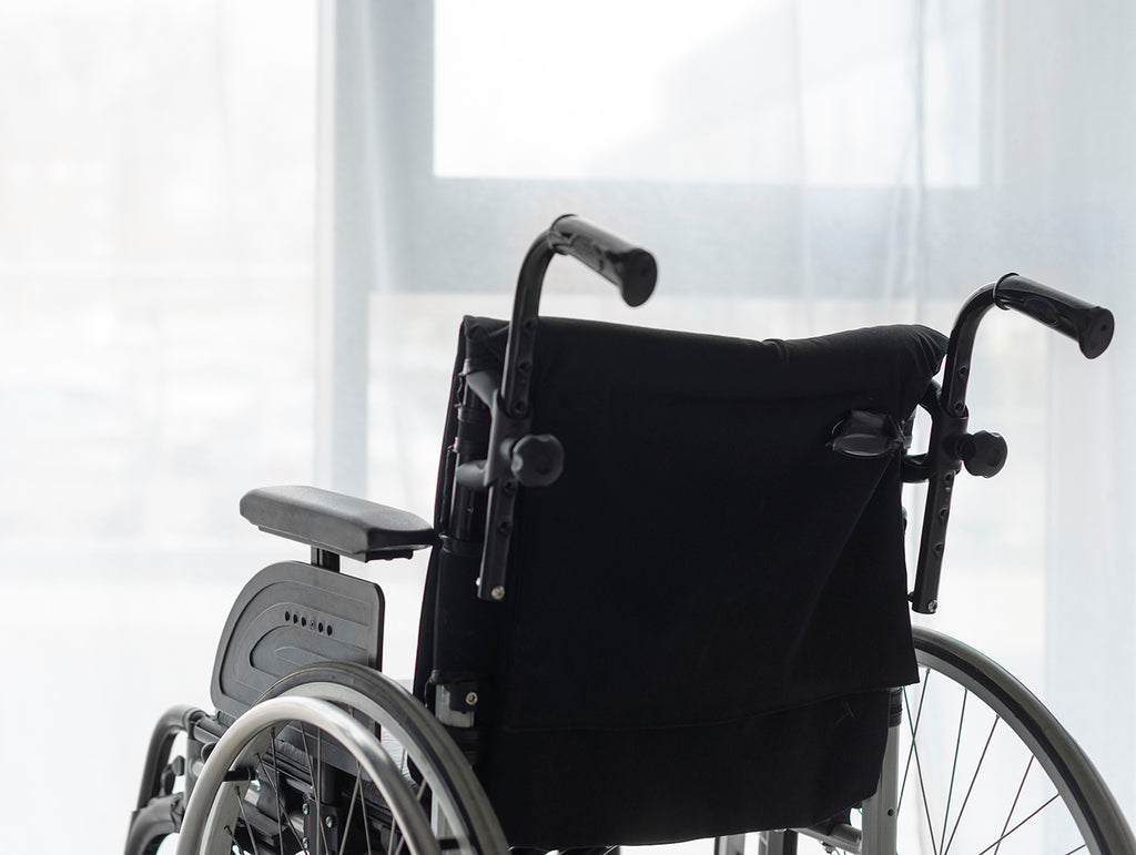 a black manual wheelchair is viewed from the back at an angle. the wheelchair is positioned in front of a plate-glass window in a well-lit room