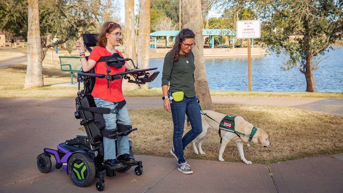 A woman in a red shirt sits in a purple and black motorized wheelchair with a headrest, being pushed by another woman in a park next to a dog with a service dog vest. The scene is set outdoors with trees, a pathway, and a body of water in the background.